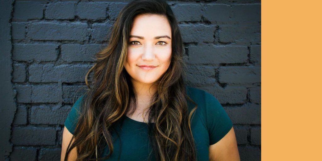 Person with long wavy hair standing in front of a dark brick wall with a mustard vertical strip, used as the hero image for Therapy for Fearful Patterns and Fear‑Response Hypnotherapy