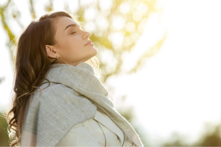 Person outdoors with eyes closed and face lifted to the light, taking a soft breath in a calm, natural setting.