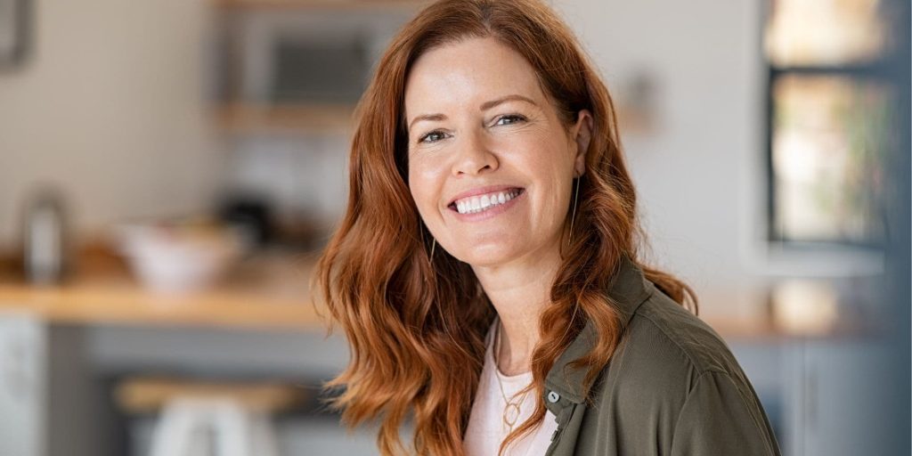Woman standing in a warm kitchen with a soft, natural smile, looking calm and more at ease after releasing fear‑based reactions.