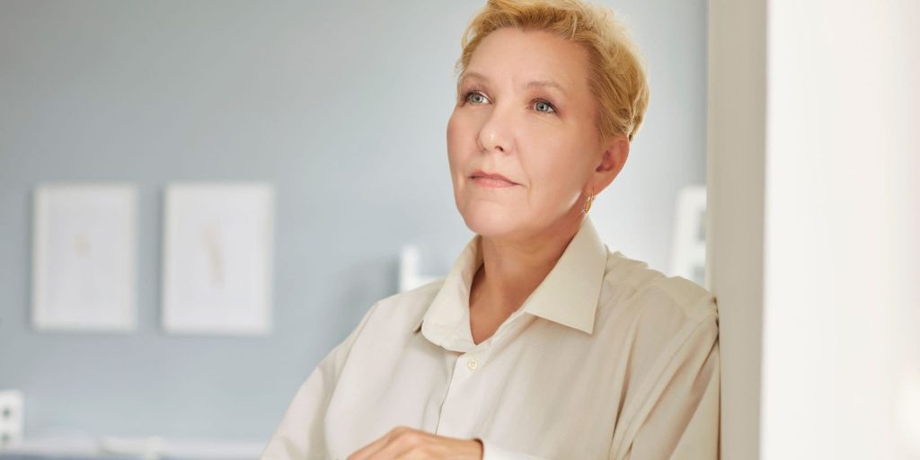 Woman standing indoors holding her glasses, looking thoughtful and calm after releasing unfair guilt and building healthier boundaries.