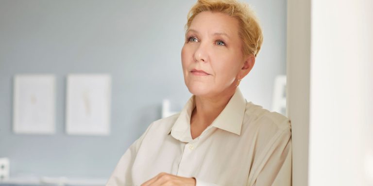 Woman standing indoors holding her glasses, looking thoughtful and calm after releasing unfair guilt and building healthier boundaries.
