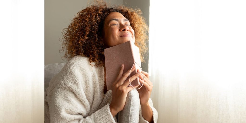 Woman wrapped in a soft blanket holding a book to her chest, looking peaceful and settled.