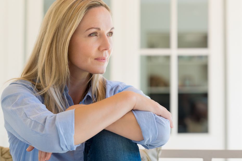 A woman sitting quietly with her arms around her knees, looking out of a window in a calm, reflective moment that represents the weight of unfair guilt and over‑responsibility.