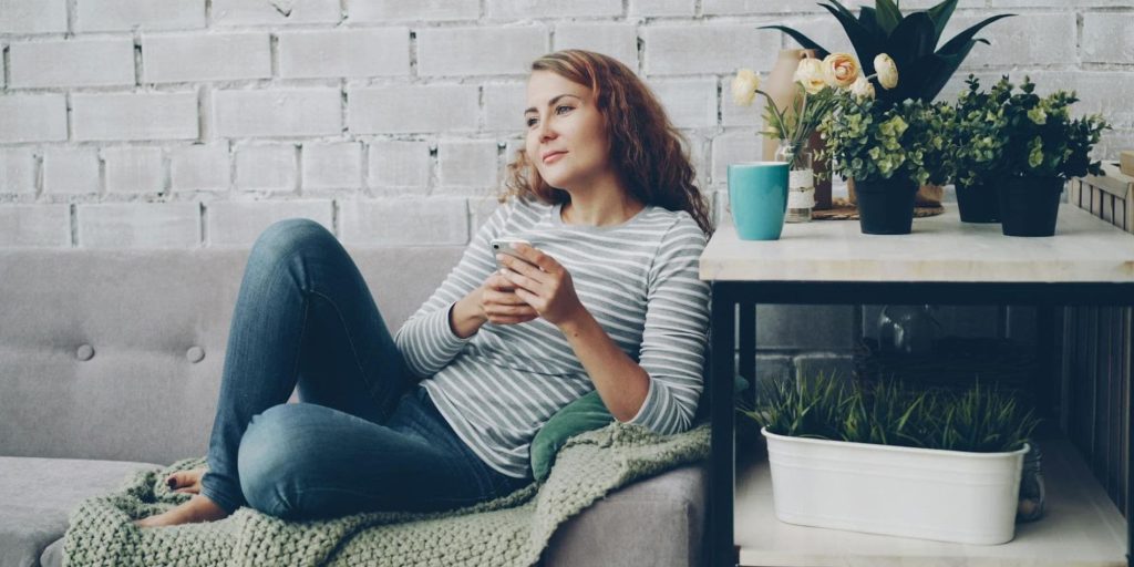 A woman sitting on a sofa in a quiet indoor setting, looking down at her phone with a calm, thoughtful expression.