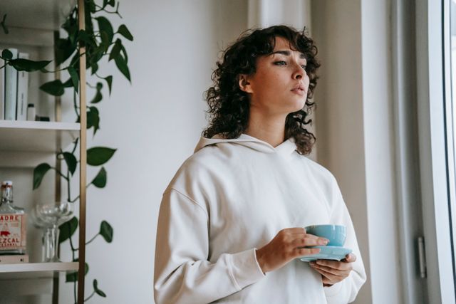 Woman holding a cup of tea by a window, looking thoughtful.